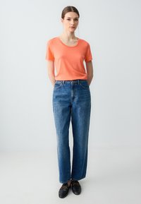 T-shirt arancione a maniche corte abbinato a jeans a gamba dritta in denim chiaro, con tasche frontali e scarpe slip-on nere con dettagli in hardware dorato.