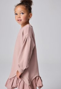 Nuori tyttö kiharat hiukset korkealla nutturalla, yllään löysä, pitkähihoinen pölyisen vaaleanpunainen mekko, jossa on röyhelöhelma, sivuprofiilissa yksinkertaisella taustalla.