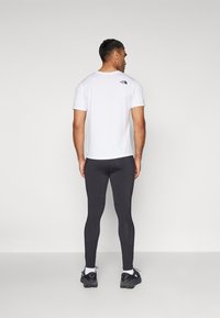 Vit t-shirt med kort ärm och en svart logotyp på övre delen av ryggen, tillsammans med figurnära svarta leggings och svarta sportskor.