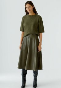 Pull en maille vert olive à manches courtes associé à une jupe midi en cuir vert olive fluide et des bottes noires à talons hauts jusqu'aux genoux.