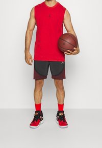 Czerwona koszulka bez rękawów, czarne i bordowe szorty z zakrzywionymi akcentami, czerwone skarpetki i czarno-czerwone buty do koszykówki. Trzyma koszykówkę.