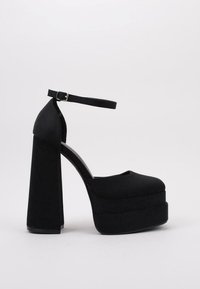 Zapato de tacón alto negro con plataforma, puntera redondeada, correa en el tobillo y un grueso tacón bloque. Fabricado con un material suave y mate.