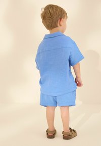 Camisa de algodón de manga corta azul claro y pantalones cortos a juego con un acabado texturizado, combinados con sandalias marrones, vistos desde la parte posterior.