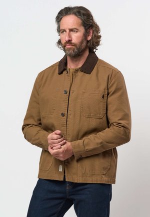 Homme aux cheveux mi-longs et barbe portant une veste marron à boutons avec un col foncé, debout devant un fond clair uni.
