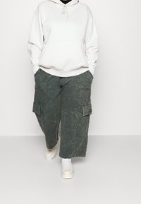 Bílá mikina s kapucí, oversize střih, spárovaná se světle zelenými cargo kalhotami s bočními kapsami a neutrálně zbarvenými botami.