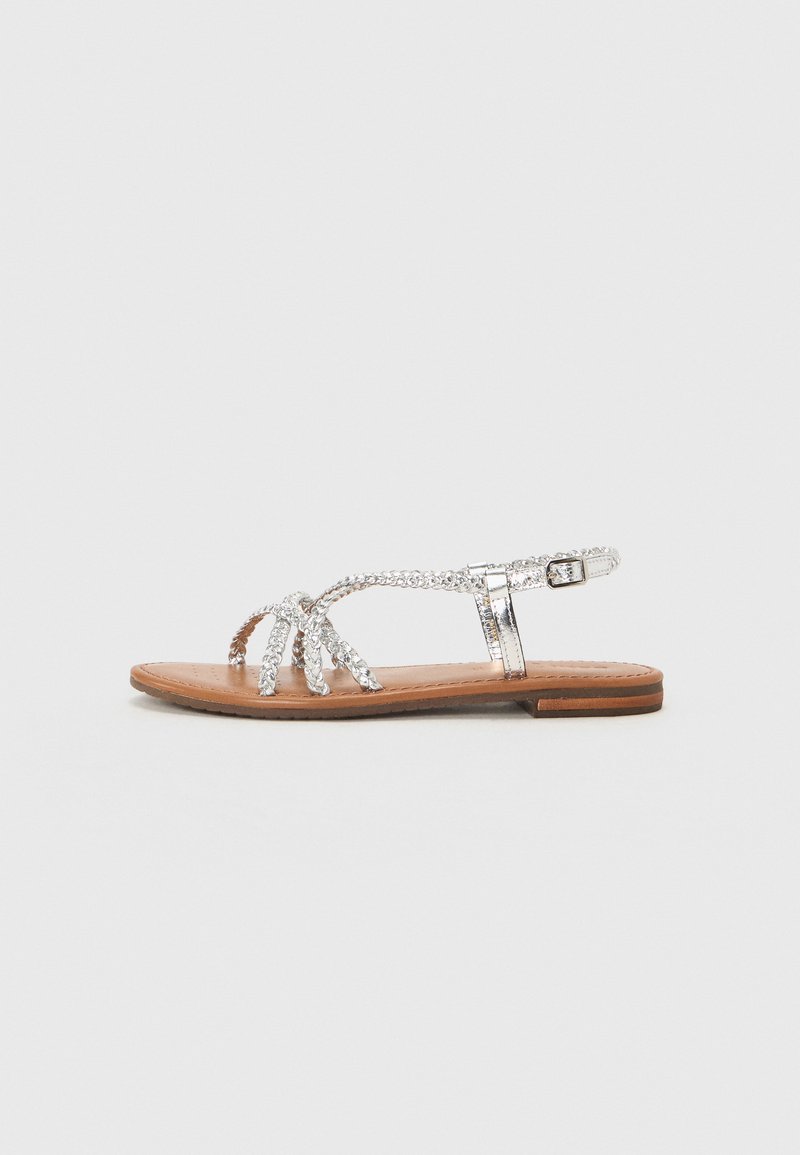 Sudraba plakanas sandales ar dekoratīvām siksnām, kas rotātas ar rhinestone akcentiem, ar brūnu ādas iekšpusi un minimālu papēdi. Regulējama potītes siksna.