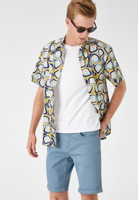 Camisa de manga corta con un patrón geométrico en amarillo, azul y marino; combinada con pantalones cortos de un azul claro; modelo lleva gafas de sol.