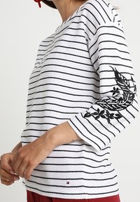 Witte trui met zwarte strepen, driekwartmouwen en zwart geborduurd bloemenmotief op de linkermouw. Inclusief logodetail.