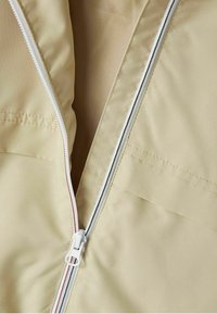 Giacca beige con zip dal tessuto liscio, dotata di una zip bianca con dettagli rossi e neri e una piccola tasca nascosta sul lato sinistro.