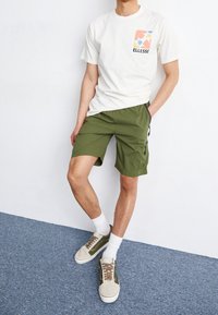 T-shirt blanc avec logo ELLESSE coloré, shorts olive avec poches latérales, et baskets gris/marron avec semelles blanches et accents contrastés.