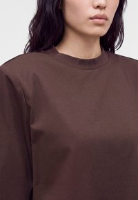 Woman wearing dark brown crew neck shirt, shown from the neck to mid-chest with shoulder-length dark hair.