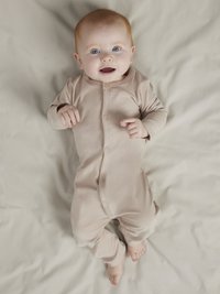 Ljusbeige babyoverall i ett stycke, långärmad, mjukt tyg, knäppning med tryckknappar framtill, resår i ärmslut och vid fötterna, ligger på en neutralfärgad yta.