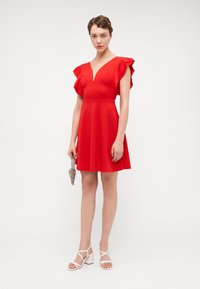 Robe rouge avec des manches courtes à volants, décolleté en V, jupe évasée, fabriquée en tissu lisse. Modèle tenant une pochette métallique, portant des sandales à talons carrés blanches.