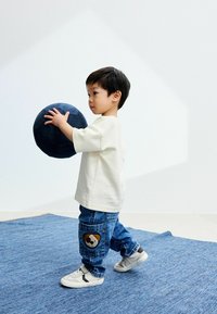 Bambino in una maglietta bianca a maniche corte e jeans di denim blu con una patch a forma di panda, tiene in mano una palla di colore blu scuro e texture, su un tappeto blu.
