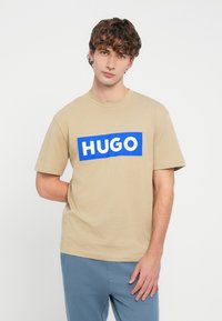Béžové bavlnené tričko s okrúhlym výstrihom, krátkymi rukávmi a veľkým modrým obdĺžnikovým grafikou s nápisom "HUGO" v bielej farbe.