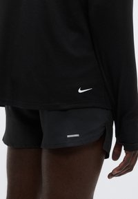 Top atlético preto de manga comprida com tecido texturizado e logótipo branco da Nike ao lado de calções pretos com um pequeno detalhe refletor perto da bainha.