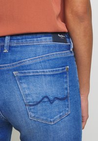 Los jeans de mezclilla azul tienen un corte de tiro medio, costuras en contraste y un bolsillo trasero con un diseño bordado ondulado. Etiqueta de la marca en negro en la cintura.