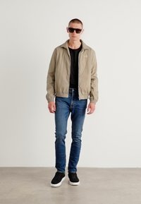 Helle beige Zip-Jacke über einem schwarzen Shirt, blaue Jeans und schwarze Sneakers mit weißen Sohlen. Das Model trägt dunkle Sonnenbrillen.