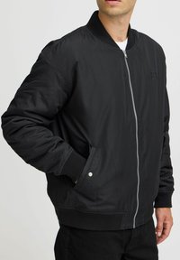 Črna bomber jakna iz gladkega materiala z sprednjim zadrgo, rebrastimi manšetami in stranskimi žepi. Ima minimalen logotip.