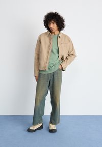 Beige canvas jacket with zip front, green sweatshirt underneath, loose blue denim pants, and beige sneakers with black soles.