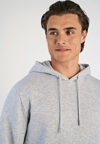 Felpa grigia con cappuccio, realizzata in tessuto morbido. Presenta lacci con tappi grigi e un design semplice, adatta per l'abbigliamento informale.