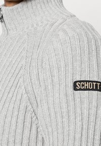 Pull en maille côtelée gris clair avec un col montant, des coutures diagonales et une étiquette noire portant le nom de la marque "SCHOTT" sur l'épaule.