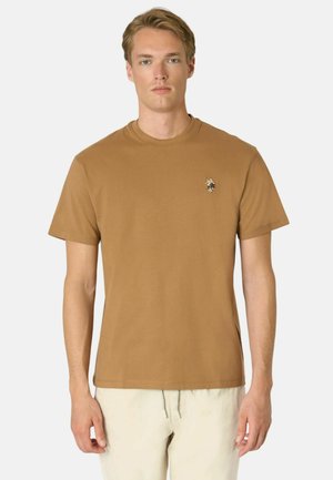 Giovane uomo che indossa una maglietta marrone tinta unita a maniche corte con un piccolo logo ricamato sul petto sinistro e pantaloni di colore chiaro.