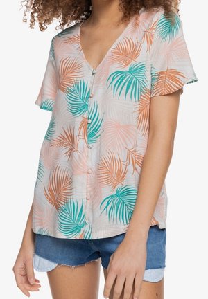Vrouw draagt een kort mouwtje blouse met knopen en een palmbladprint in blauwgroen, perzik en bruin, gecombineerd met versleten spijkershorts.