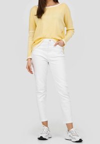 Keltainen neulepaita pitkillä hihoilla, valkoisilla yksityiskohdilla, yhdistettynä valkoisiin slim-fit-farkkuihin ja harmaisiin sekä valkoisiin tennareihin. Kokovartalokuvanäkymä.