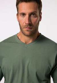 Camiseta verde oliva de cuello en v con una textura suave, corte relajado y detalles de costura mínima, llevada por un modelo masculino con una expresión neutral.