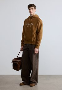 Brun hoodie med frontficka och vit text "CALVIN", kombinerad med vida bruna byxor, håller en brun läderväska och bär beige skor.