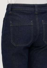 Närbild av en mörkblå denimjeans bakficka och söm på en person som bär en vit topp.