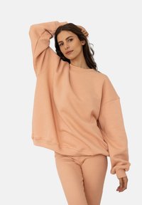 Przesadny sweter w kolorze brzoskwiniowym z prążkowanymi mankietami i okrągłym dekoltem, w połączeniu z dopasowanymi spodniami o wąskim kroju. Miękka tekstura, minimalistyczny design.