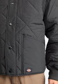 Jachetă neagră matlasată cu nasturi tip clapetă, buzunar mare în față, manșete cu nervuri și o mică etichetă cu logo-ul Dickies deasupra tivului.