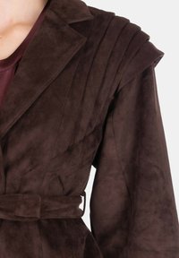 Giacca in camoscio marrone con design su misura, ampi revers e spalle plissettate, chiusa con una cintura coordinata in vita.