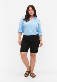 Camisa de botones azul claro con mangas enrolladas, combinada con pantalones cortos negros y sandalias marrones. Tejido suave con un detalle de bolsillo.