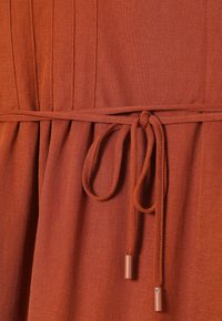 Tissu de couleur rouille avec ceinture nouée en forme de nœud à la taille, présentant des détails de coutures verticales et un matériau tissé texturé.