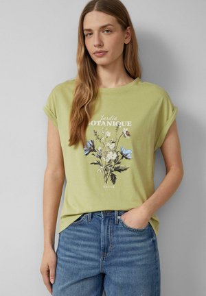 Kobieta z długimi jasnobrązowymi włosami, ubrana w jasnooliwkowy T-shirt z podwiniętymi rękawami i kwiatowym nadrukiem oraz niebieskie dżinsy, stojąca na tle jednolitego szarego tła.