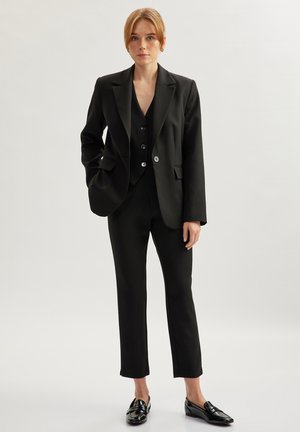 Black tailored suit featuring a blazer with sharp lapels, a matching vest with button accents, and slim-fit trousers. Glossy black shoes complete the look.