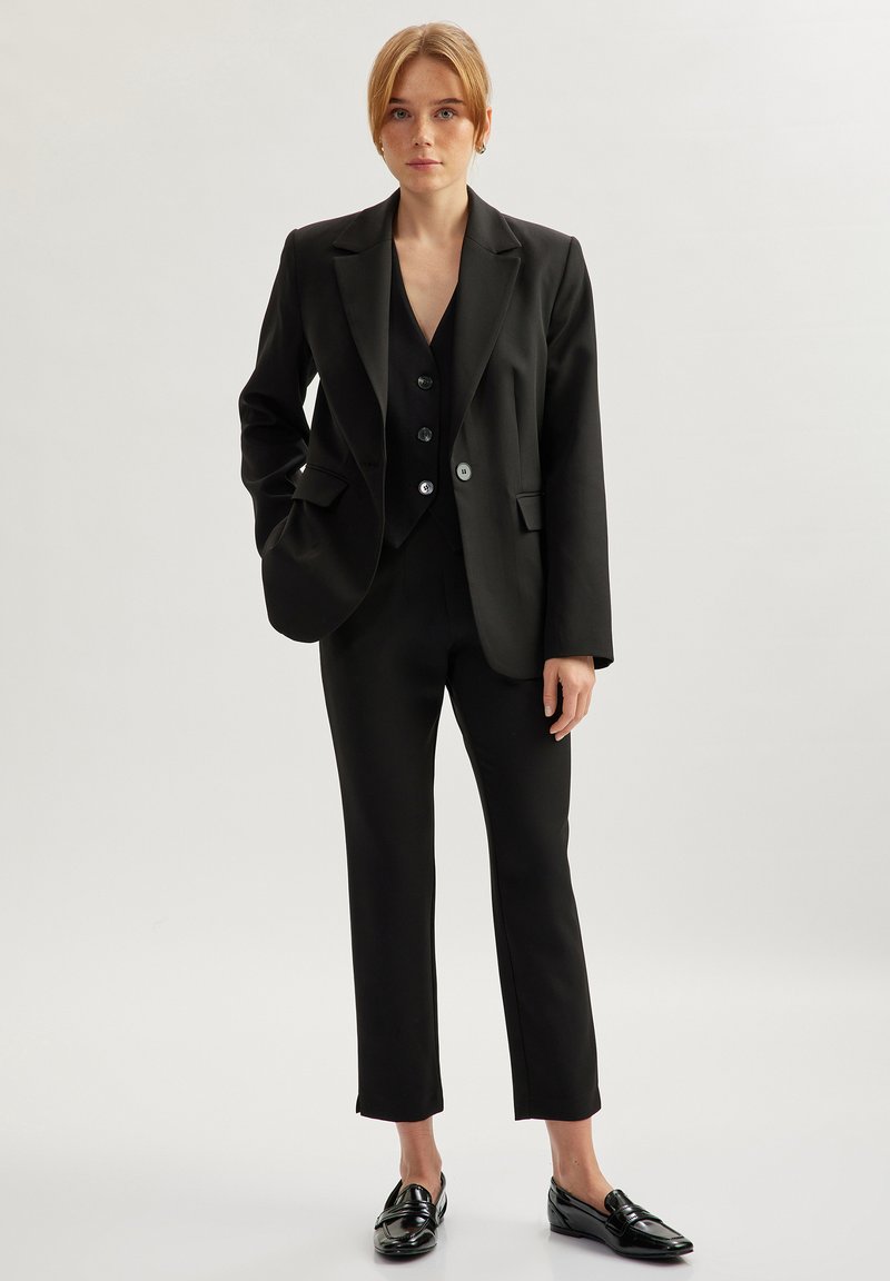 Zwart op maat gemaakt pak met een blazer met scherpe revers, een bijpassend vest met knoopaccenten en slim-fit broek. Glanzende zwarte schoenen maken de look compleet.