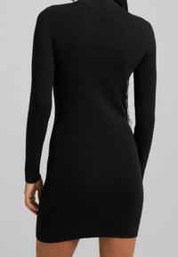 Svart ribbad klänning med långa ärmar, figursydd design och hög halsringning. Har en slät textur och längd över knäna.