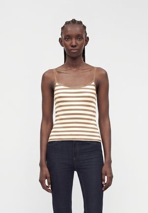 Femme aux cheveux tressés portant un haut à fines bretelles beige et blanc à rayures et un jean bleu foncé, debout devant un fond uni.
