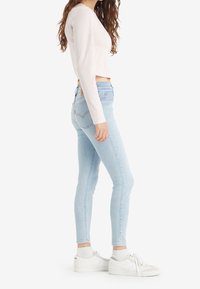 Woman in light blue skinny jeans, white long-sleeve top, white socks, and white sneakers, standing sideways against a plain background.