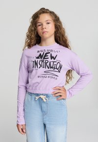 Lawendowa koszula z długim rękawem z elastycznym dołem i graficznym nadrukiem, zestawiona z jasnoniebieskimi dżinsami z elastycznym paskiem.