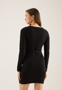 Robe noire à manches longues, avec un motif en losanges texturé et une taille ceinturée. Design ajusté, tissu lisse et coupe au genou.