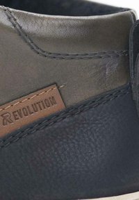 Lederschuh mit dunkelgrünen und schwarzen Texturen, einem braunen Etikett mit "REVOLUTION" und sichtbaren Nahtdetails entlang der Kanten.