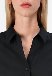 Camicetta nera con colletto e chiusura a bottoni, caratterizzata da una texture liscia e un design dritto. Inquadratura ravvicinata che mette in risalto il collo e i dettagli della camicia.