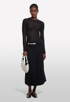 Femme portant un haut noir transparent à manches longues, une jupe midi noire, des chaussures pointues noires, tenant un sac à main blanc clouté avec une ceinture blanche.
