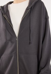 Tamno siva zip-up dukserica s kapuljačom na vezanje, prednjim džepovima i rebrastim manžetama. Napravljena od mekog materijala s glatkom teksturom.