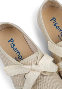 Beige canvas slip-on shoes with a textured finish, featuring a decorative bow and an embossed logo on a beige insole.